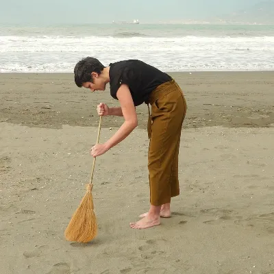 Vemos a Jana barriendo la playa. Con una escoba entre sus manos, va vestida con pantalón marrón y una camiseta negra. Tiene el pelo corto. De fondo, vemos el mar.