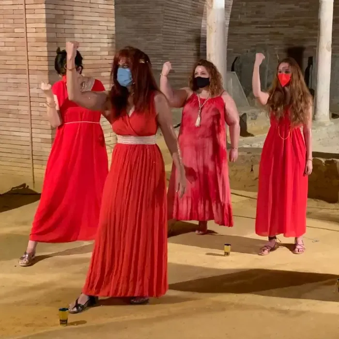Cuatro mujeres con vestidos largos rojos miran al frente; llevan mascarillas y sandalias. Se encuentran en el interior de Museo de Arte Romano de Mérida. Con su mano derecha, alzan su puño en señal de resistencia y fuerza.
