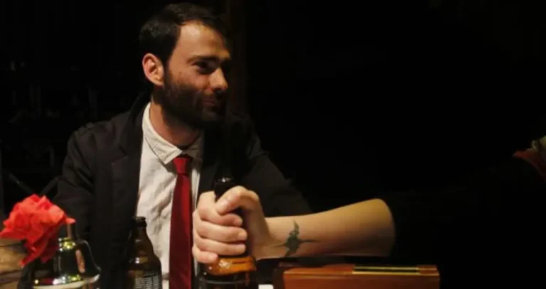 Un hombre vestido con smoking negro, camisa blanca y corbata roja está sentado en una mesa dentro de una habitación. Se encuentra mirando a su derecha a una persona a la que no podemos ver más que su brazo extendido que agarra una botella de una bebida alcohólica.