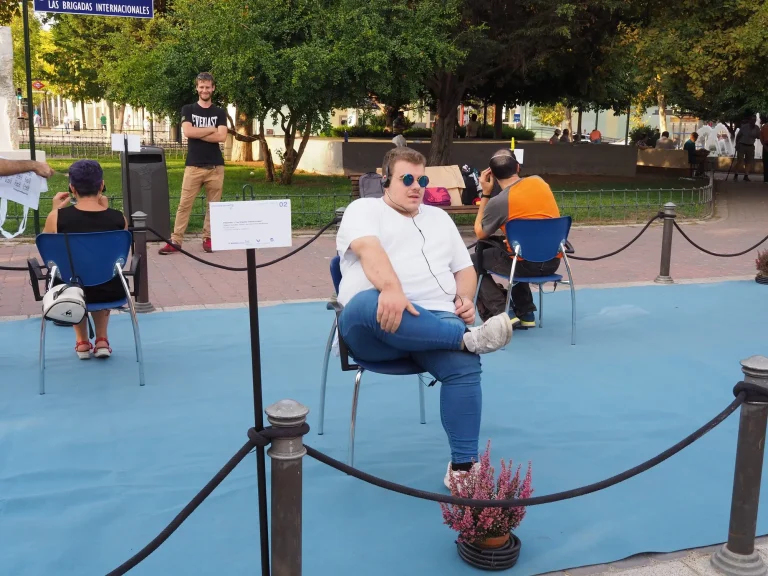 Vemos a un hombre sentado en una silla en un exterior. Lleva gafas de sol, una camiseta de manga corta y unos pantalones vaqueros. Está escuchando algo que se reproduce a través de los auriculares. Hay más personas alrededor sentadas y escuchando. El suelo está cubierto con una gran alfombra azul.