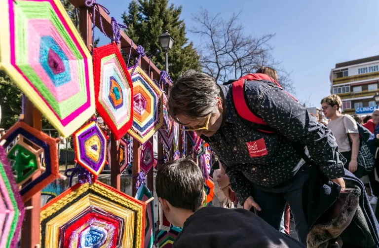 Estamos en el exterior de un día soleado. En unos barrotes, quedan colgadas unas estructuras de tela de colores que se han tejido manualmente. Un hombre y un joven las miran con curiosidad.