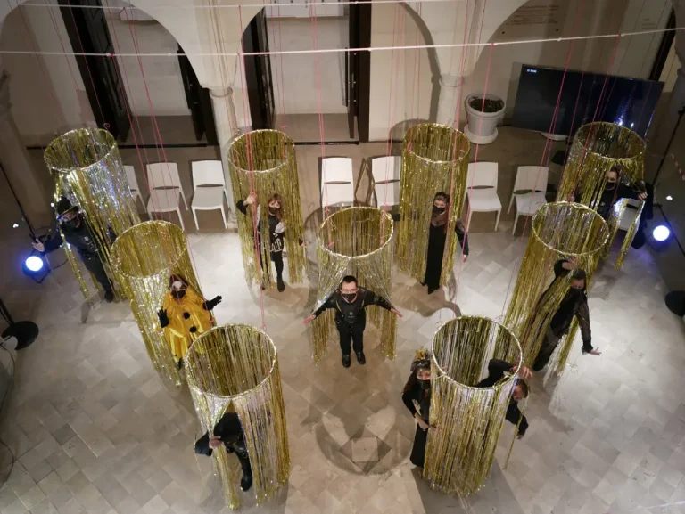 Desde el cielo, vemos unas esferas doradas que esconden a los/as participantes de la performance. A excepción de una persona que va vestida de amarillo, todas van de negro y con mascarilla negra.