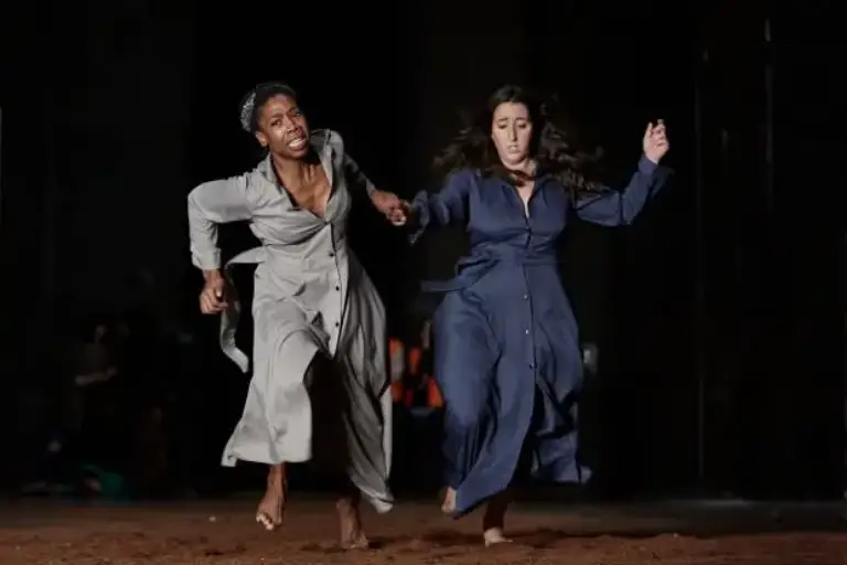 Dos mujeres huyen de la playa de noche sin mirar atrás. La de la derecha, viste un traje color gris en contraste con su piel negra. La de la derecha, viste un vestido azul oscuro en contraste con su piel blanca.