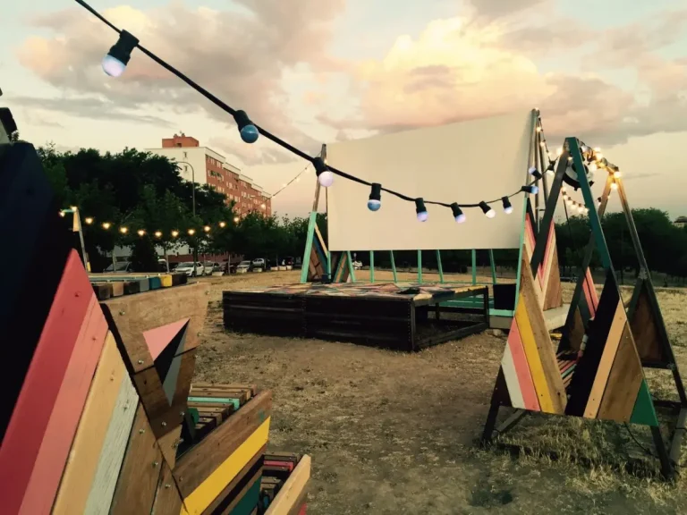 Un atardecer en el cine de verano. Vemos una pantalla blanca al fondo con un pequeño escenario delante. Alrededor, columpios con luces colgadas en ellos rodean e iluminan el lugar.