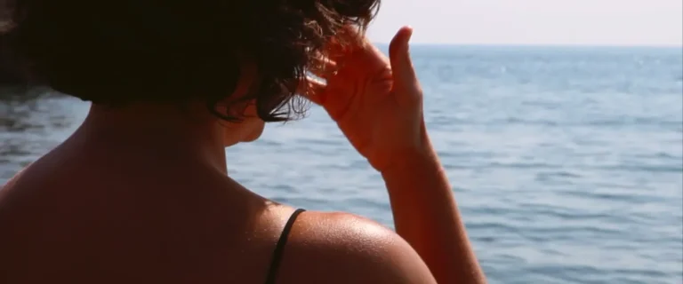 Jana está de frente al mar, se toca la frente con los dedos de su mano derecha. Asoma por su hombro un tirante de color negro.