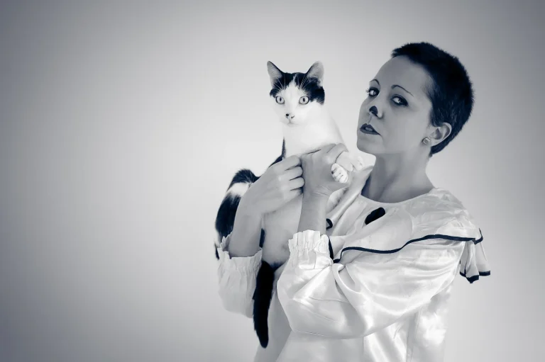 Una foto en blanco y negro. Vemos a Jana vestida de payaso blanco. Sobre sus brazos, a la altura de su cabeza, sostiene a su gato Shiva de color blanco y negro. Tiene los labios pintados de color negro y en la nariz ha replicado la mancha negra que también tiene Shiva.
