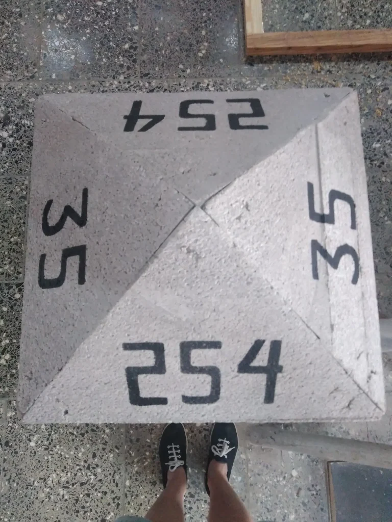 Vemos desde un plano cenital una estructura piramidal blanca. En cada una de sus caras, vemos nº escritos en negro. En la parte inferior de la foto, asoman unas piernas con zapatillas negras que están curioseando.