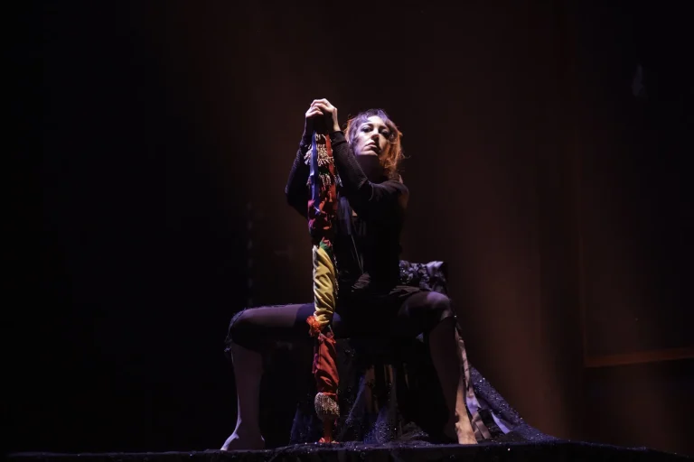 Una mujer en un escenario, se encuentra sentada en un taburete con las piernas abiertas y en medio sujetando una bandera enroscada a su propio mástil. La mujer va vestida de negro y mira al frente con la barbilla alzada, como orgullosa y desafiante.
