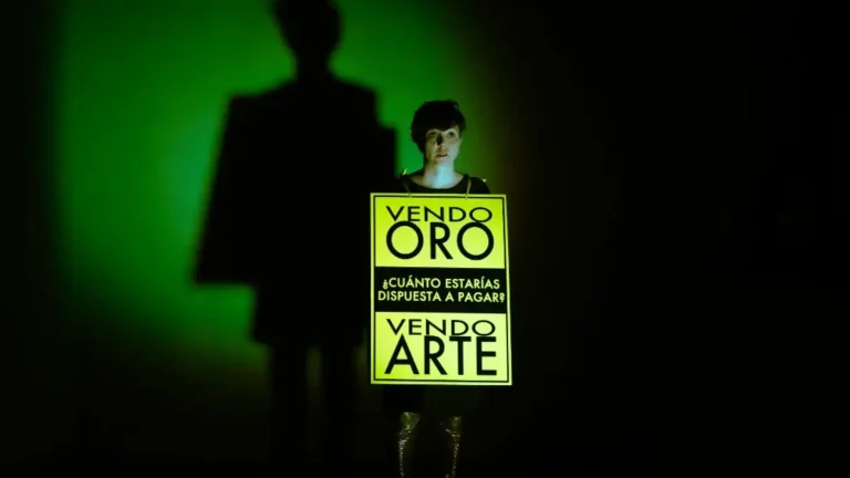 Jana se encuentra en una habitación oscura con un foco que la ilumina de frente. Lleva colgado sobre sus hombros un cartel que indica "Vendo oro. ¿Cuánto estarías dispuesta a pagar? Vengo arte." Las letras están escritas en negro sobre fondo amarillo.