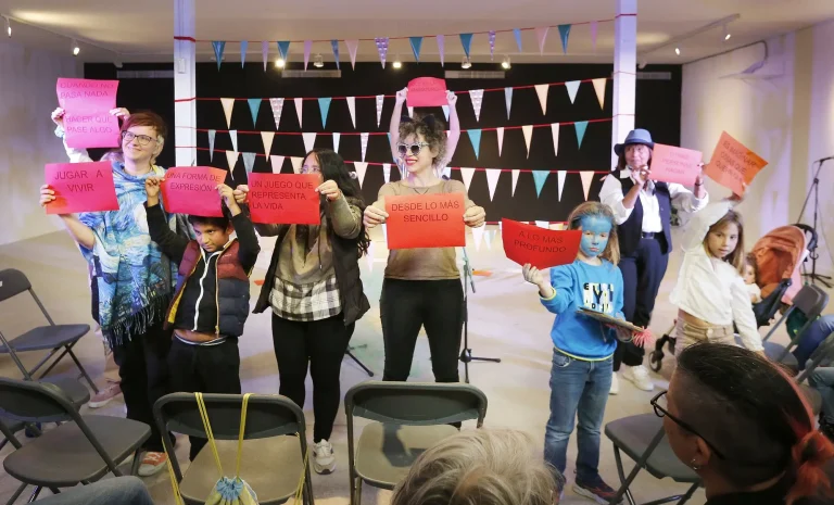 Estamos en una sala. Varias personas y niñas/os sujetan carteles de colores con mensajes y los enseñan al público que está sentado en sillas. Al fondo, vemos entre dos columnas, colgados unos banderines de colores.