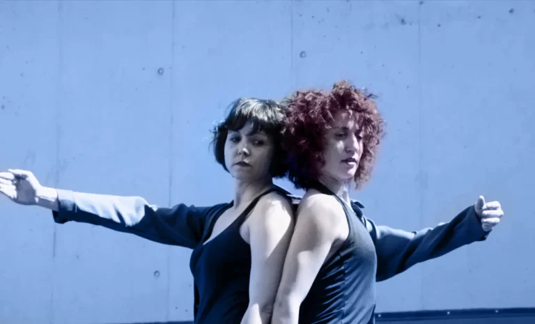 Dos mujeres se encuentran unidas por una americana. A la izquierda, Jana extiende su brazo derecho a la horizontal y su compañera, pelirroja y de pelo rizado, extiende también su brazo izquierdo a la misma altura. Ambas van vestidas con una camiseta de tirantes negra que deja al descubierto los brazos que no están en la americana.