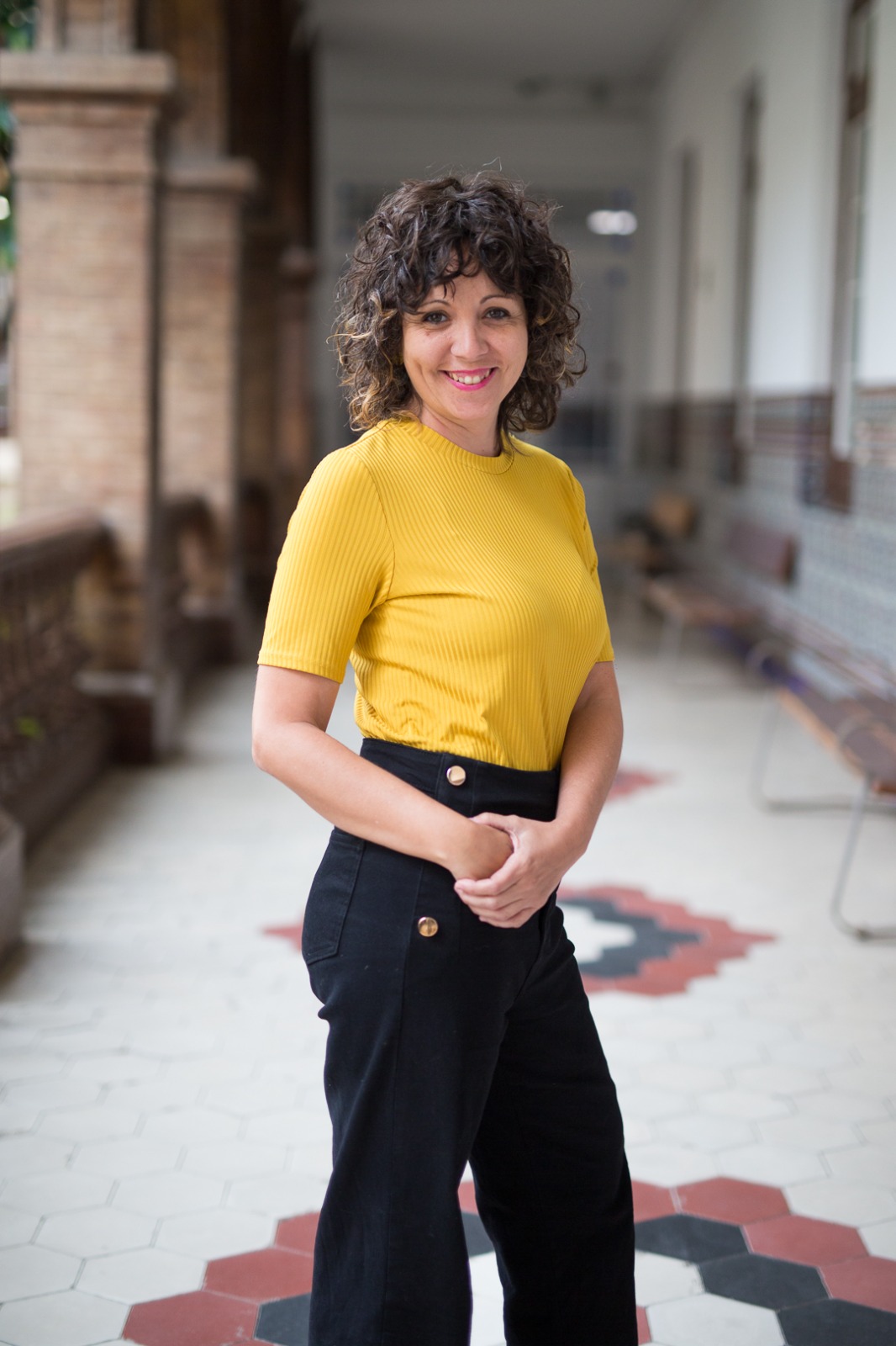 Vemos a Jana posando a cámara en uno de los pasillos exteriores de La Térmica. Sonríe y sujeta sus manos a la altura de su cintura. Lleva una camiseta amarilla y un pantalón negro.